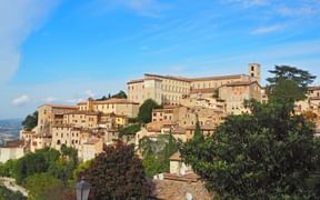 Hügelstadt Todi in Italien mit Steingebäuden, Glockenturm und Kloster auf der Spitze. Grüne Bäume rahmen die mittelalterliche Architektur unter blauem Himmel.