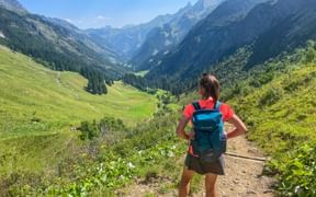 Wanderin mit blauem Rucksack auf Bergweg mit Blick auf das grüne Stillebachtal in den Allgäuer-Lechtaler Alpen mit Gipfeln in der Ferne.