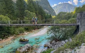 Two hikers crossing a suspension bridge over turquoise Soča River in Trenta, Julian Alps. Mountains and forest visible in background.