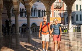 Two hikers with backpacks walking through a sunlit colonnade with stone arches in Udine, Italy. Historic buildings with arched windows visible.