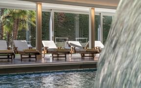 Innenpool-Bereich im Hotel Adria Meran mit Holzliegen, Wasserspiel und Glaswänden mit Blick auf tropische Pflanzen und efeubedeckte Wände.