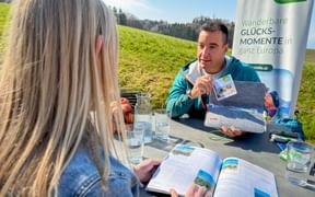 Eurohike-Berater zeigt einer Frau Reisebroschüren an einem Tisch im Freien mit grüner Wiese und Waldhintergrund. Banner zeigt 'Glücksmomente'.