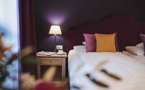 Hotelzimmer mit weißer Bettwäsche, lila und orangen Kissen, Holznachttisch mit Lampe und lila Vorhängen im Hotel Adria Meran.