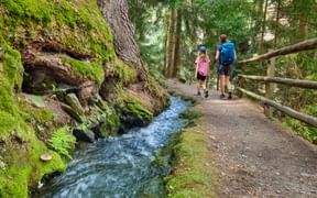 Two hikers walking on a forest path beside a flowing stream in Schluderns, South Tyrol. Moss-covered rocks and wooden railings line the trail.
