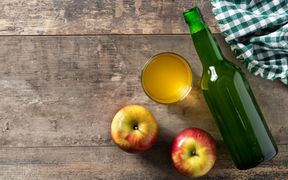 Zwei Äpfel, ein Glas mit Apfelessig, eine Flasche mit Apfelessig, ein grün-weiß-kariertes Geschirrtuch auf einer Holzplatte