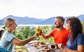 Three hikers toasting with beer at a wooden table with food, overlooking Lake Tegernsee and mountains in Bavaria.