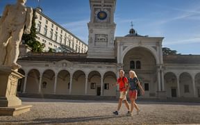 Zwei Wanderer überqueren die Piazza Libertà in Udine mit Uhrturm, Arkadengang und klassischer Statue unter blauem Himmel.