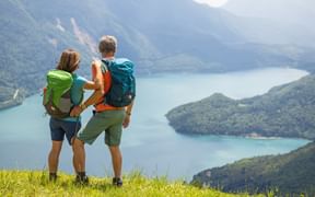Two hikers with backpacks standing on grassy hillside overlooking Lake Molveno surrounded by forested mountains in the Italian Alps.