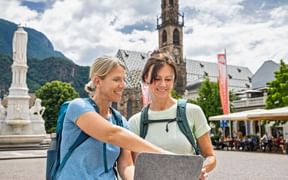 Zwei Frauen mit Rucksäcken betrachten ein Tablet am Waltherplatz in Bozen. Hinter ihnen ein weißes Denkmal, Kirchturm und Berge.