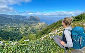 Female hiker with blue backpack sitting among white hydrangea flowers, overlooking Madeira's dramatic north coast landscape with mountains and ocean.