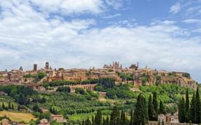 Panoramablick auf Orvieto auf einem Vulkanfelsen, umgeben von Zypressen und grünen Hügeln unter blauem Himmel mit weißen Wolken.