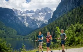 Three hikers with trekking poles walking along Gosausee lake in Salzkammergut. Dachstein glacier mountain and forested slopes in background.