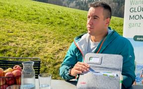 Man in teal jacket presenting gray travel bag at outdoor table with water carafe and apples, green meadow and forest in background.