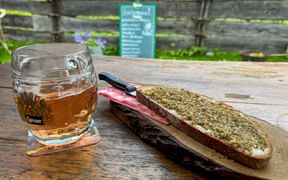 Glass of beer and bread with herb spread on wooden board at mountain hut. Rustic wooden fence and menu board visible in background.