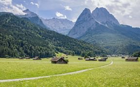 Grüne Wiese mit Weg und Holzhütten in der Region Garmisch. Bewaldete Berge und felsige Zugspitzgipfel unter blauem Himmel mit weißen Wolken.