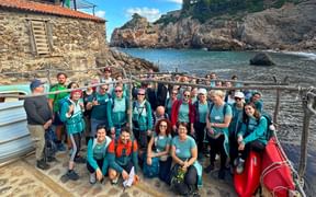 Große Gruppe von Wanderern in türkisfarbenen Shirts posiert an einer felsigen Küstenbucht auf Mallorca mit Steingebäuden und Klippen im Hintergrund.