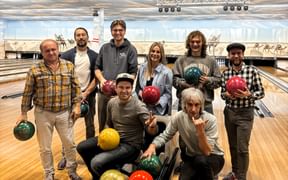 Gruppe von acht Personen posiert mit bunten Bowlingkugeln in einer Bowlingbahn und lächelt in die Kamera. Bowlingbahnen im Hintergrund sichtbar.