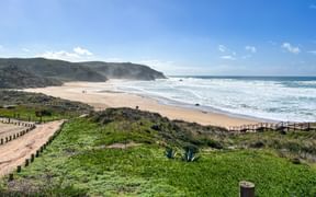 Panoramablick auf den Strand Praia do Amado mit goldenem Sand, weißen Wellen und dramatischen Klippen. Holzstege durchqueren grüne Küstenvegetation.