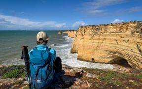 Wanderer mit blauem Rucksack und Kappe sitzt am Klippenrand der Algarve und blickt auf goldene Kalksteinklippen und türkises Meer.