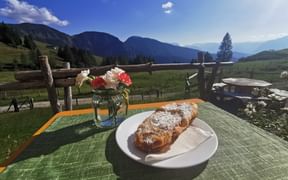 Powdered sugar-dusted Pofesen on white plate with flowers in vase, wooden fence and mountain landscape near Hintersee in background.