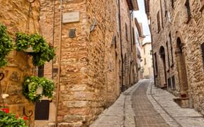 Enge Kopfsteinpflasterstraße in Spello mit Steingebäuden. Ein Schild mit 'Via Povera Vita' ist an der Wand mit blühenden Pflanzen sichtbar.