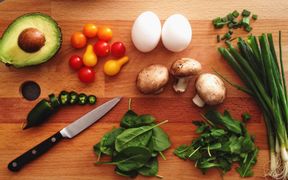 Avocado, Tomaten, Eier, Pilze, Frühlingszwiebel, Spinat, Ruccola und eine grüne Chili auf einem Schneidbrett mit einem Messer