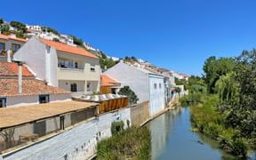 Weiße Häuser mit roten Ziegeldächern in Aljezur entlang eines ruhigen Wasserlaufs. Das Hügeldorf zeigt traditionelle portugiesische Architektur unter klarem blauen Himmel.