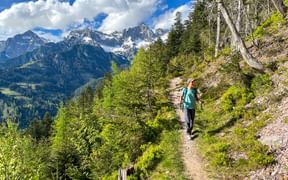 Wanderer mit Rucksack auf Bergweg im Hollersbachtal, Hohe Tauern. Schneebedeckte Gipfel und grüne Wälder unter blauem Himmel mit weißen Wolken.