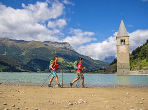 Hikers on the shore of Lake Reschen with a view of the church tower