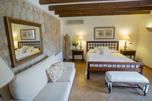 Geräumiges Doppelzimmer mit Steinwand, Holzbalken, weißem Sofa und dunklem Holzbett. Terrakottafliesen bedecken den Boden.