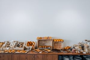 Frühstücksbuffet mit frischem Gebäck, Brötchen, Donuts und Müsli auf Holzsideboard mit gestapelten Tellern im Praia Hotel Porto Covo.