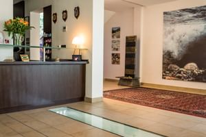 Moderne Hotelrezeption mit dunklem Holztresen, Blumen und Lampe. Beiger Fliesenboden mit Glaspaneel, Orientteppich und großem Seefoto an der Wand.