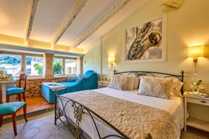 Helles Doppelzimmer mit schmiedeeisernem Bett, Holzbalken, Steinwänden, türkisfarbenem Sessel und Kunstwerk über dem Bett im Son Sant Jordi Pollenca.