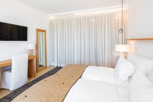 Modernes Hotel Avenida Doppelzimmer mit zwei Einzelbetten, weißer Bettwäsche, beigem Läufer, Wandfernseher und bodenlangen Vorhängen.