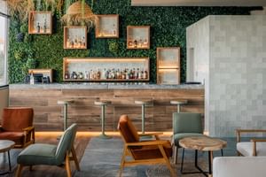 Zeitgenössische Bar im Praia Hotel Porto Covo mit vertikaler Gartenwand, Holztheke, Barhockern und Loungebereich mit orangen und grünen Sesseln.