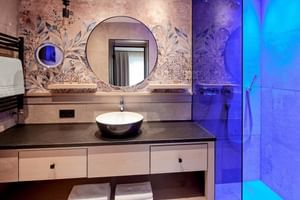 Elegantes Badezimmer mit Blumentapete, rundem Spiegel, Aufsatzwaschbecken auf dunkler Platte und blau beleuchteter Dusche im Hotel Alte Post Fieberbrunn.