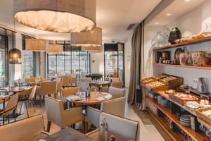 Moderner Frühstücksraum im Hotel Avenida Lagos mit beigen Stühlen, Holztischen, Buffetregalen mit Brot und Gebäck und großen Fenstern.
