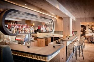 Moderne Bar im Hotel Alte Post Fieberbrunn mit geschwungenem Metallregal, Holzbalken, Bartresen mit Hockern und umfangreicher Flaschenauslage.