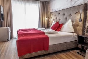 Modern double room with tufted headboard, red throw blanket, white bedding, wooden floor, and large windows with beige curtains.