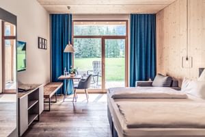 Bright hotel room at Lechzeit Elmen with wooden ceiling, twin beds, blue curtains, and glass door opening to terrace with alpine meadow view.