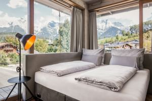 Modern double room with large windows overlooking Alpine mountains in Maria Alm. Gray bed, floor lamp, and panoramic mountain views.