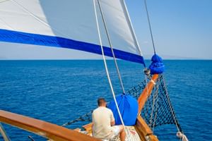 View from MS Sagena's bow showing a passenger sitting on deck under white and blue sails, with calm blue sea and clear sky in background.