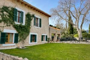 Steinfassade des Hotels Son Palou Orient mit grünen Fensterläden, efeubedeckten Wänden, Terrasse mit Stühlen und gepflegtem Rasen unter blauem Himmel.