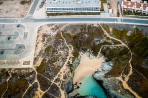 Luftaufnahme des Praia Hotel & Spa Porto Covo mit weißem modernem Gebäude, Parkplatz und abgeschiedener Strandbucht mit türkisfarbenem Wasser.