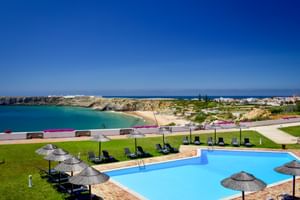 Außenpool der Pousada Sagres mit Liegestühlen und Sonnenschirmen auf grüner Wiese, mit Blick auf Sandstrand und türkisfarbenes Meer unter blauem Himmel.