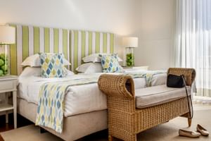 Helles Hotelzimmer mit zwei Einzelbetten, grün gestreiftem Kopfteil, geometrisch gemusterten Kissen, Rattanbank am Fußende und weißen Vorhängen.