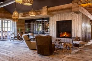 Modern alpine lounge at Sepp Hotel Maria Alm with rustic wood paneling, woven pendant lights, tan armchairs, and lit fireplace beneath wall-mounted TV.