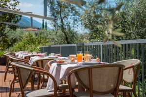 Außenterrasse mit gedeckten Frühstückstischen mit Saft und Kaffee. Rattanstühle, weiße Tischdecken und Blick auf Olivenbäume und Berge.
