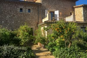 Steingebäude von Can Quatre in Sóller mit Gartenweg durch Orangenbäume voller Früchte und mediterranen Pflanzen unter blauem Himmel.