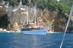 White sailing yacht MS Dalmatino anchored in blue waters near limestone cliffs covered with green vegetation in a Croatian bay.